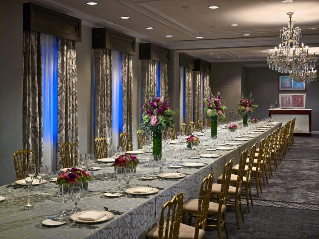 A long, elegant banquet table set with plates, glasses, and floral centerpieces is arranged in a well-lit room with chandeliers and gold chairs.