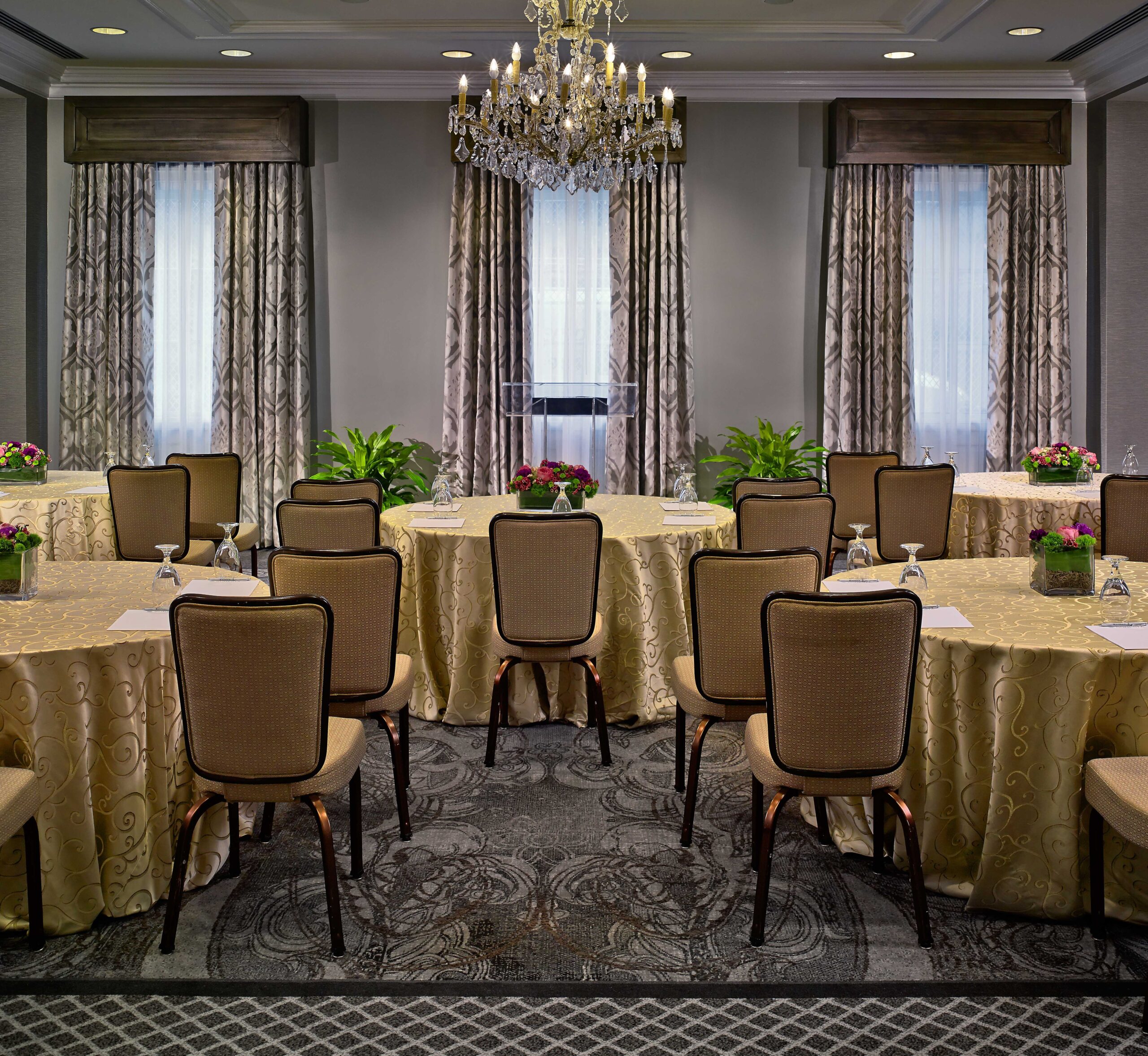 Conference room with tables arranged in a U-shape, beige chairs, floral centerpieces, notepads, water glasses, and a chandelier overhead.
