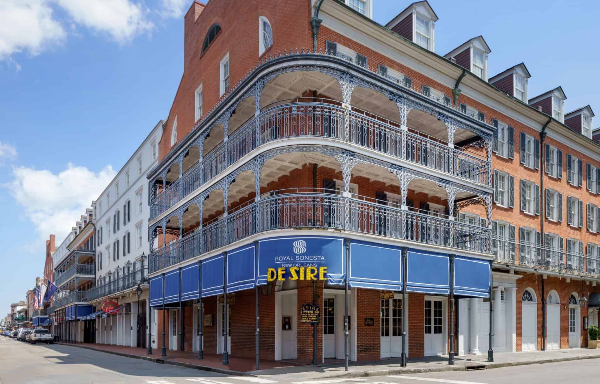 Exterior view of the Royal Sonesta New Orleans hotel, featuring classic French Quarter architecture with red brick walls, wrought-iron balconies, under a clear blue sky.